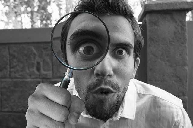 Man holding a magnifying glass up to his eye, humorously enlarging it, and expressing surprise or curiosity.