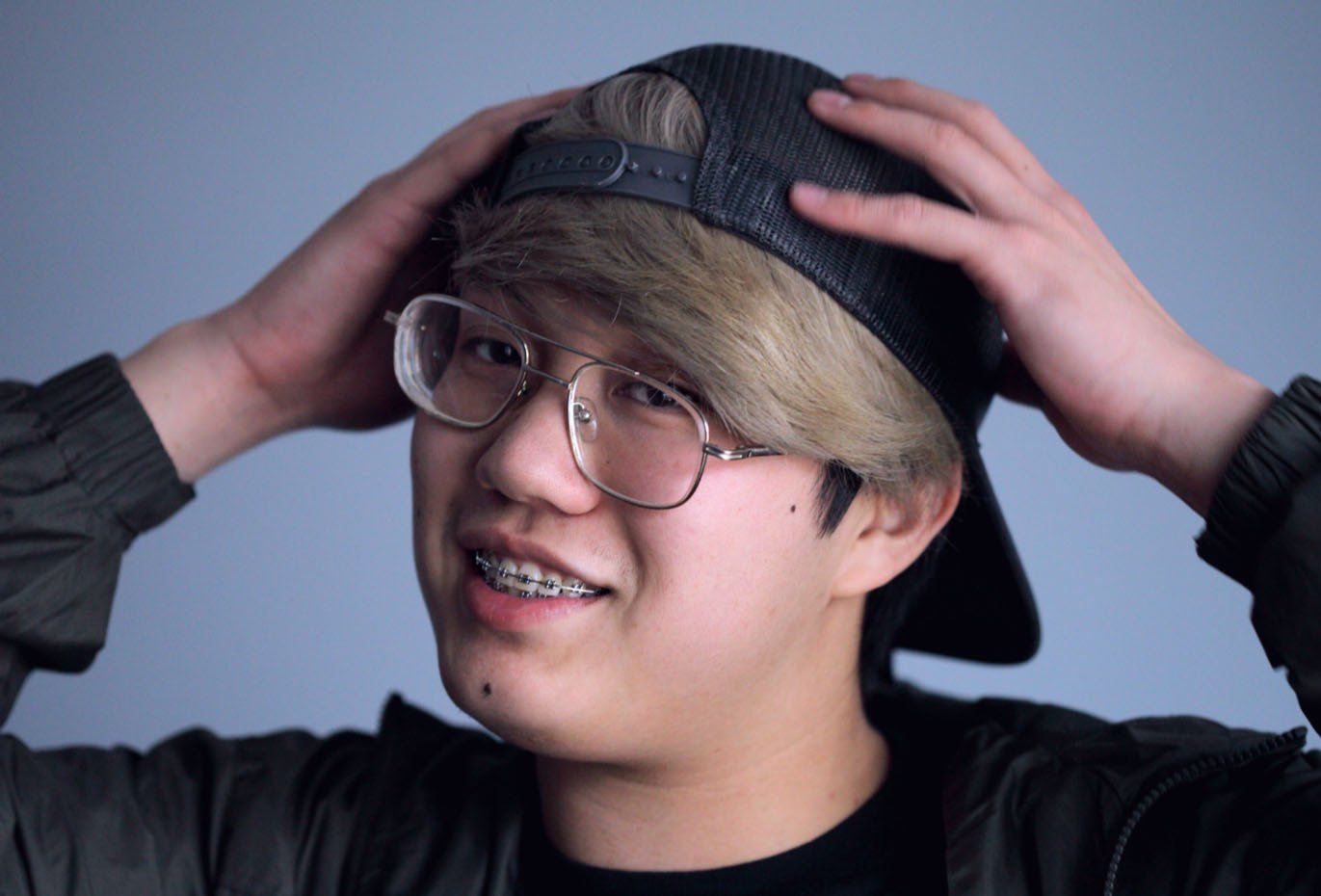 A young man with braces on his teeth is wearing a hat and glasses