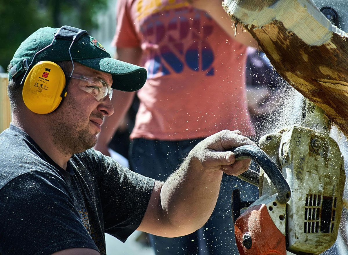 A man wearing ear muffs and goggles is using a chainsaw to cut a piece of wood.