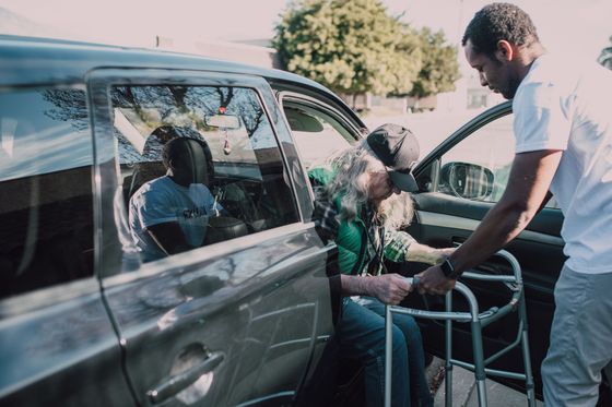 Man helping a seated person into a wheelchair beside a gray car outdoors