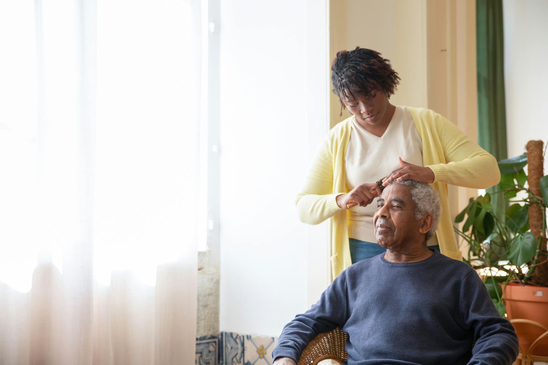 Caregiver adjusting an older man’s hearing aid beside a bright window