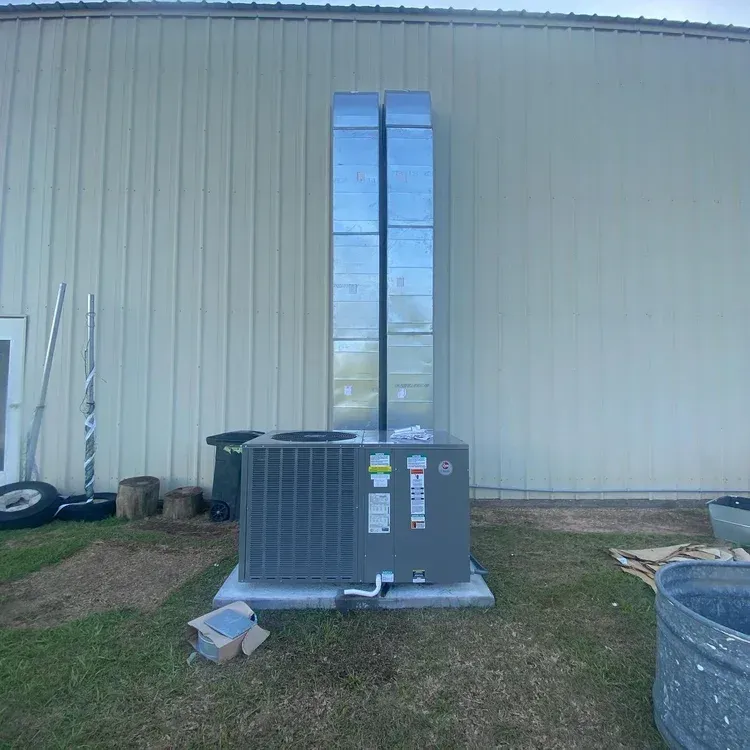 Outdoor HVAC unit beside a beige metal building with a tall silver vent duct and grass foreground