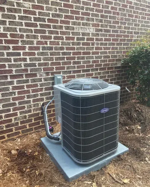 Outdoor air conditioner unit on a concrete pad beside a brick wall and shrubs