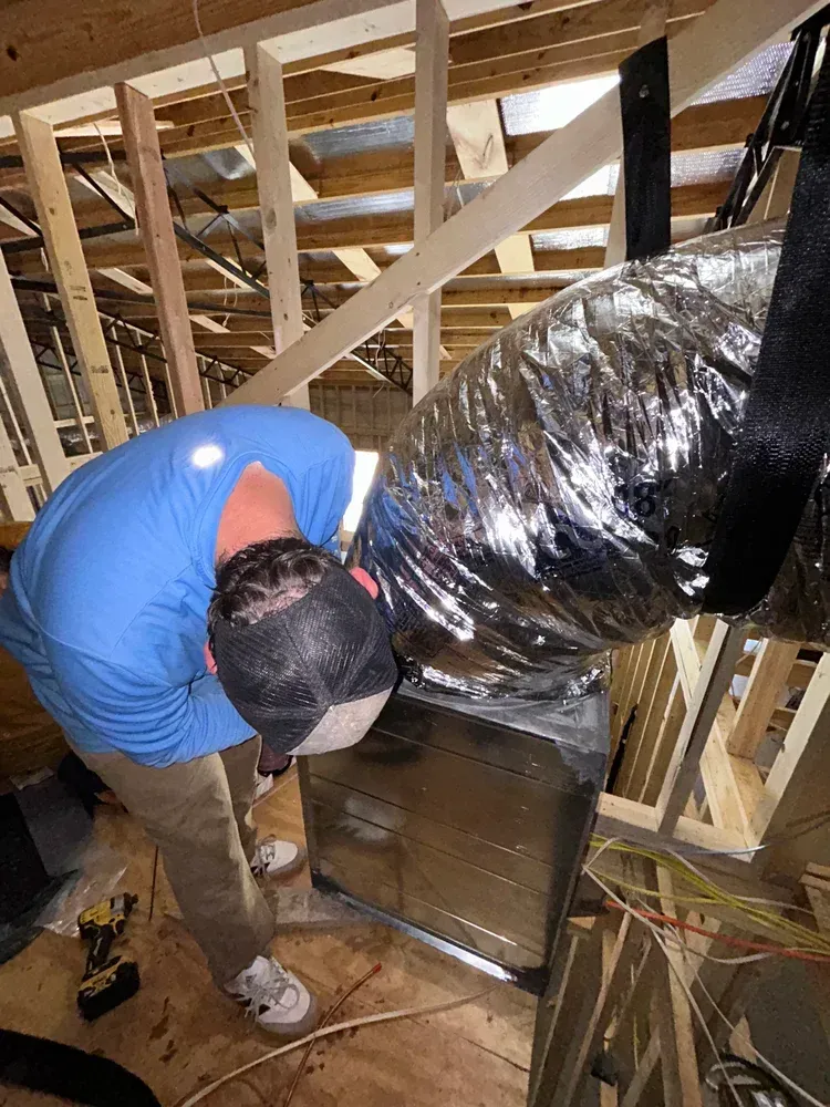Worker in blue shirt and cap inspects attic ductwork inside exposed wooden framing.