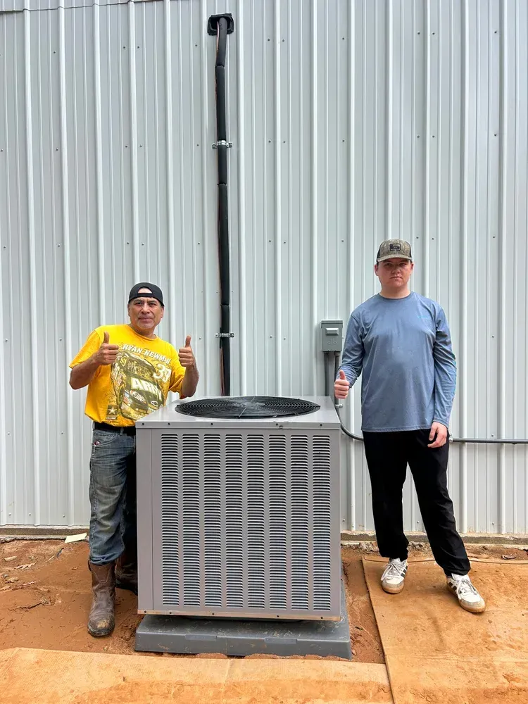 Two people stand beside a large outdoor air conditioner unit against a metal wall.