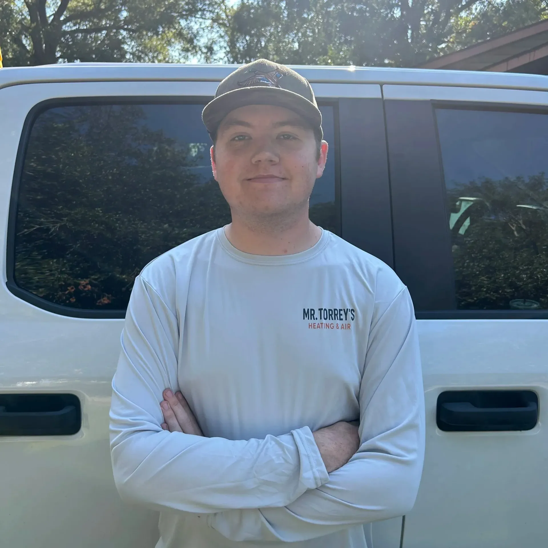 Person standing with arms crossed in front of a white van, wearing a light shirt and dark cap.