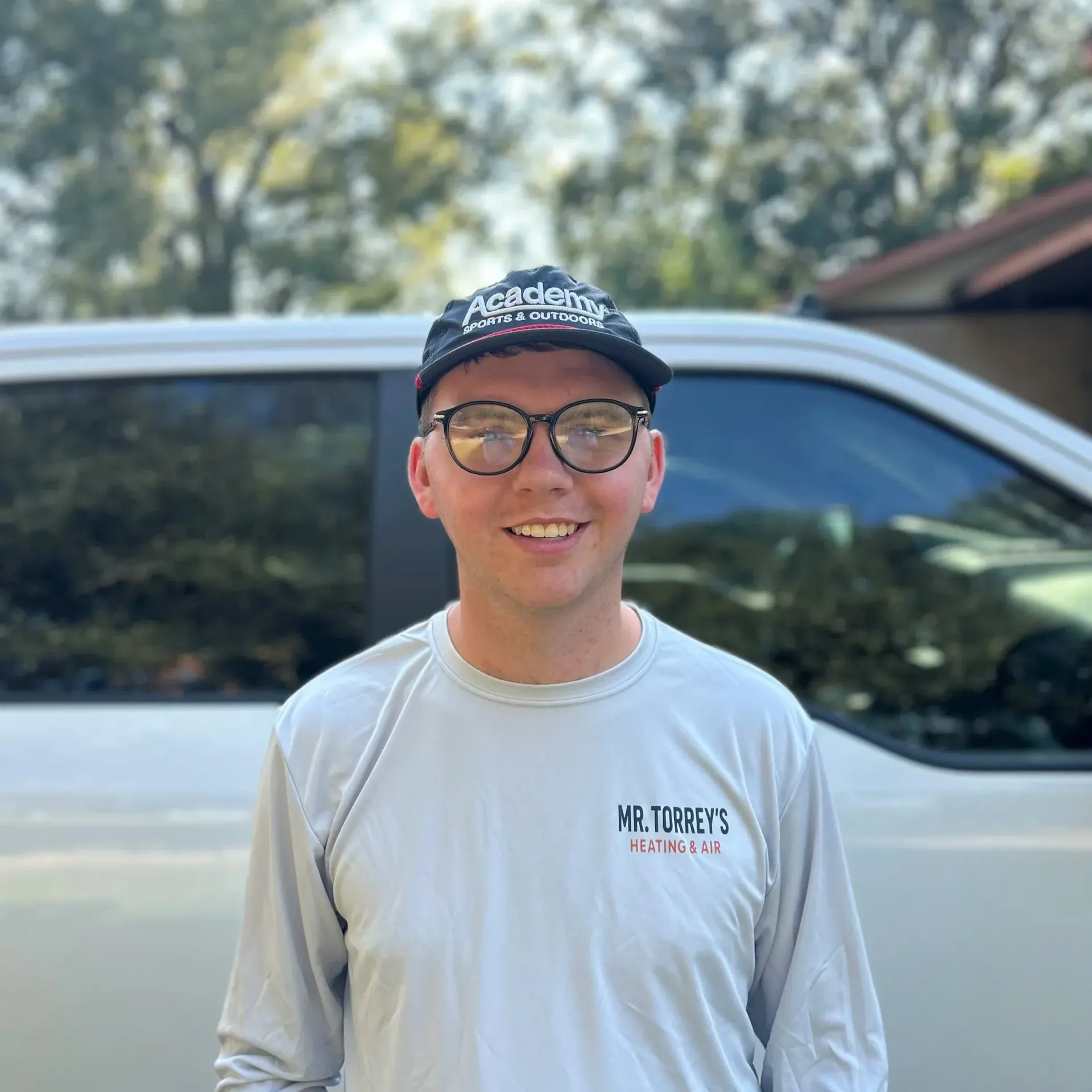 Person smiling in glasses and cap, standing outdoors beside a silver vehicle.