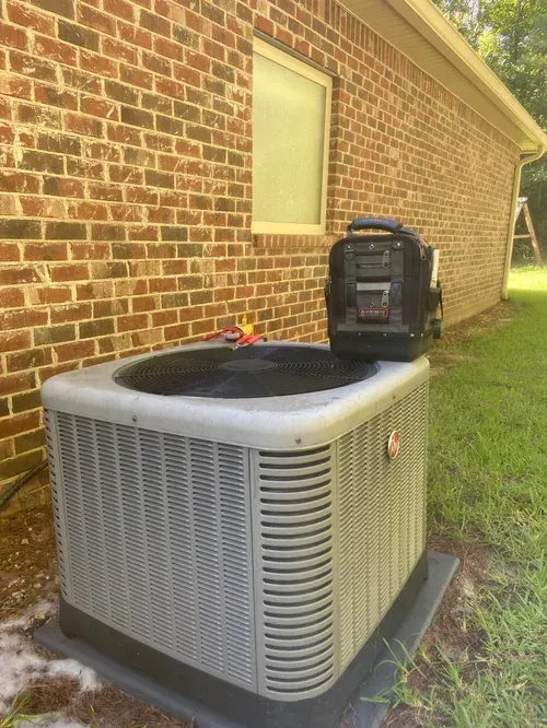 Outdoor HVAC unit beside a brick house wall and lawn, with a portable generator on top.