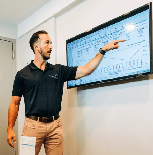 Man points at chart on wall-mounted screen. He wears black shirt, khaki pants, indoors.
