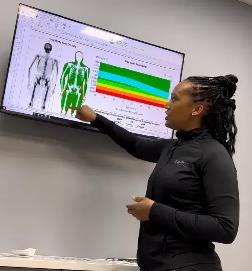 Man points to diagram on screen while another looks on, inside a brightly lit room.