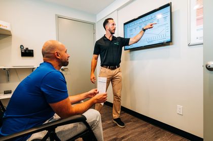 Man points at screen, explaining information to seated person in an office setting.