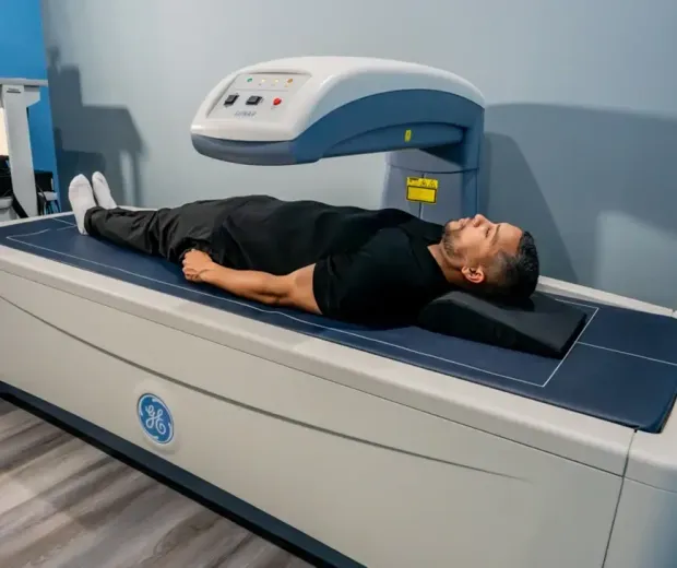 Man lying on a bone density scanner. He wears black clothing and looks up. Room with gray walls.