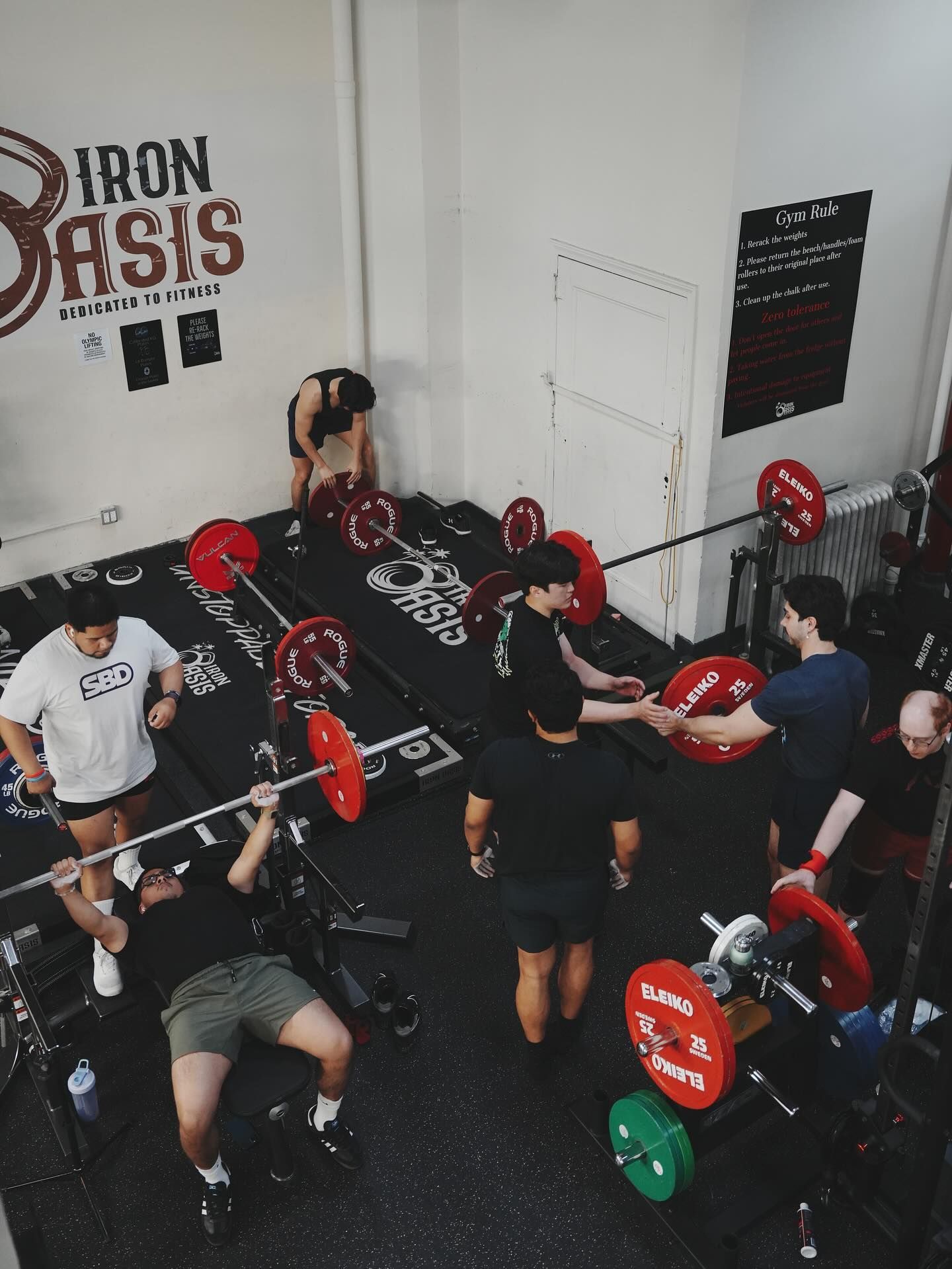 People lifting weights in a gym. Barbell bench press in progress, spotters assist. Red, green, and black weights.