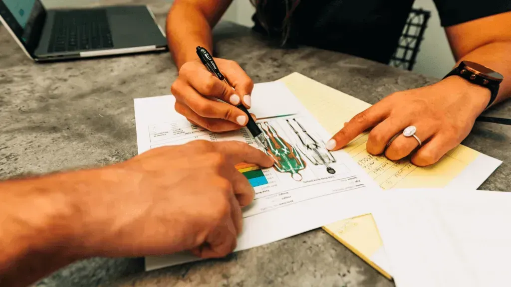 Man points to diagram on screen while another looks on, inside a brightly lit room.