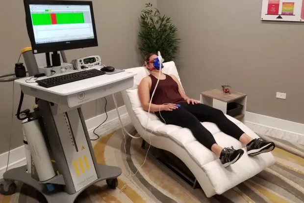 Woman undergoing metabolic testing, wearing a mask, resting on a reclined chair, monitor displays data.