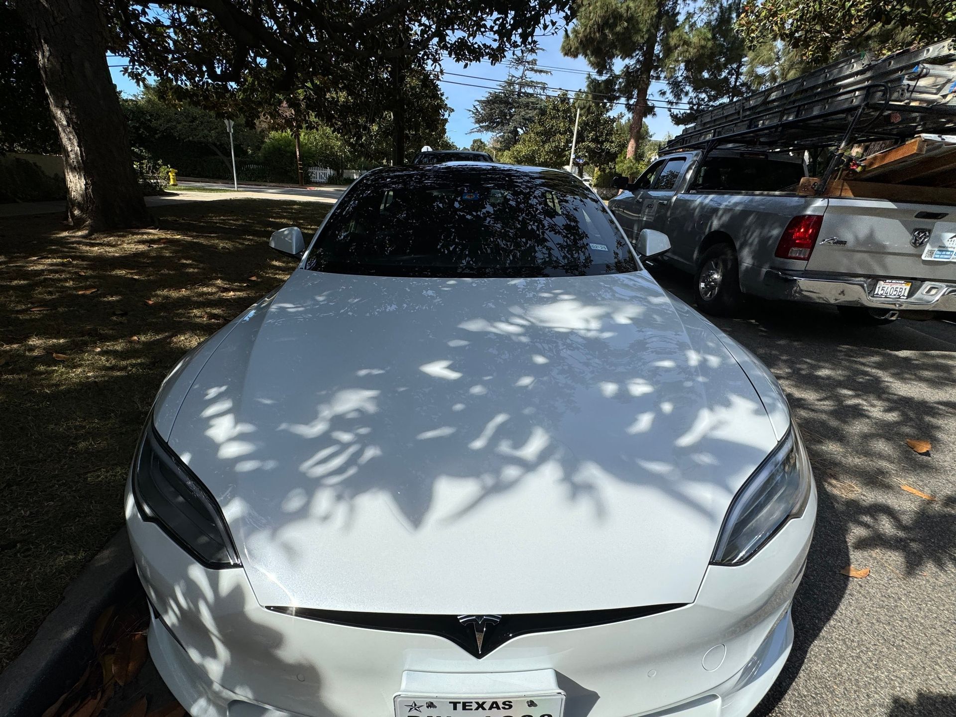 White Tesla car parked in front of a silver pickup truck on a tree-lined street on a sunny day.
