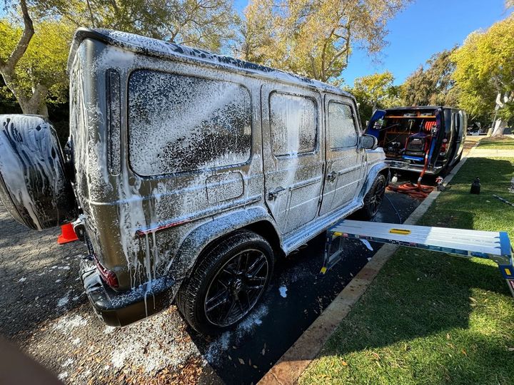Mercedes-Benz G-Wagon covered in white foam, being washed outdoors, near a grassy area and trees.
