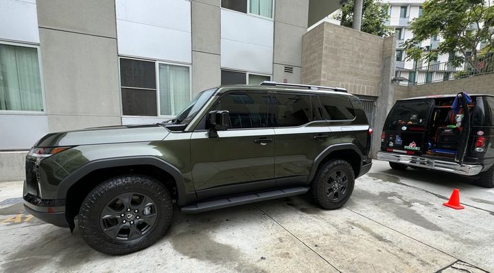 Green SUV parked next to a black van with orange cone in front, near a building.