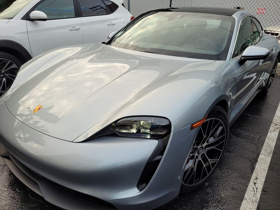 Silver Porsche Taycan electric car parked, wet asphalt, black wheels, in a parking lot.