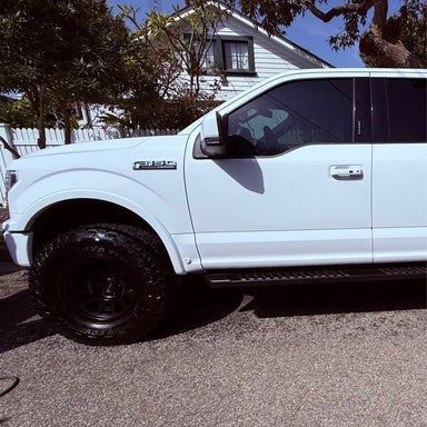 White Ford pickup truck with black wheels parked on a street. House in the background.