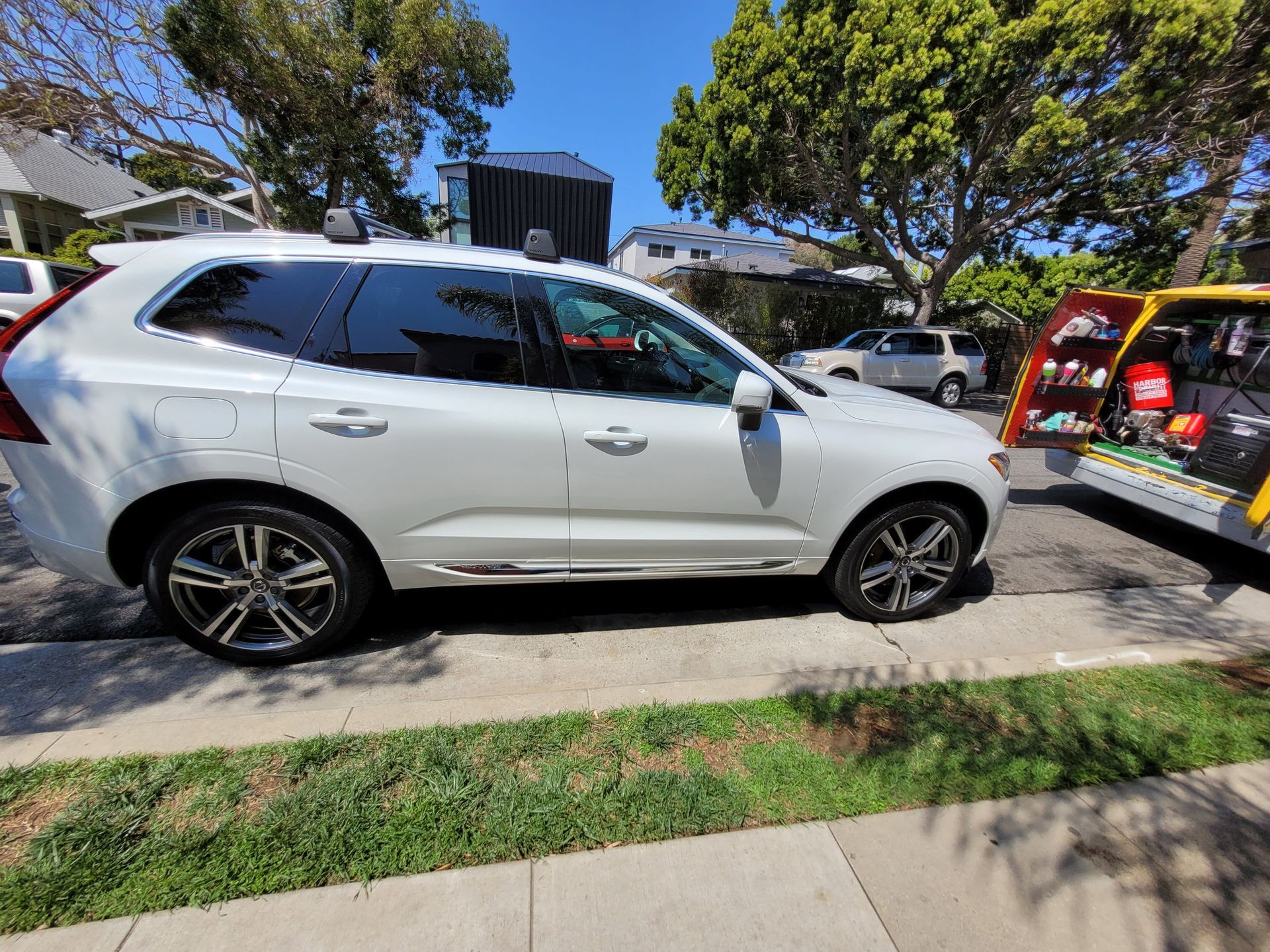 White Volvo SUV parked on a street next to a yellow service truck on a sunny day.