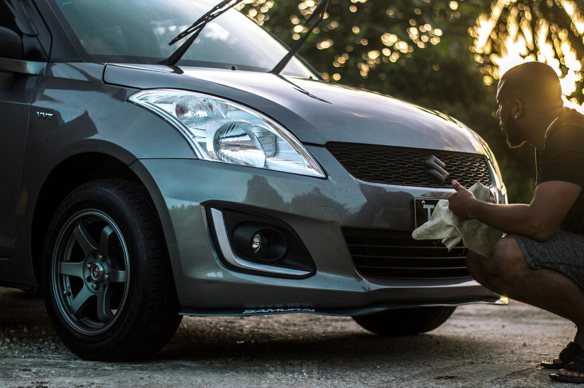 Man wiping a gray Suzuki car's front bumper. Sunlight shines on the car and man.