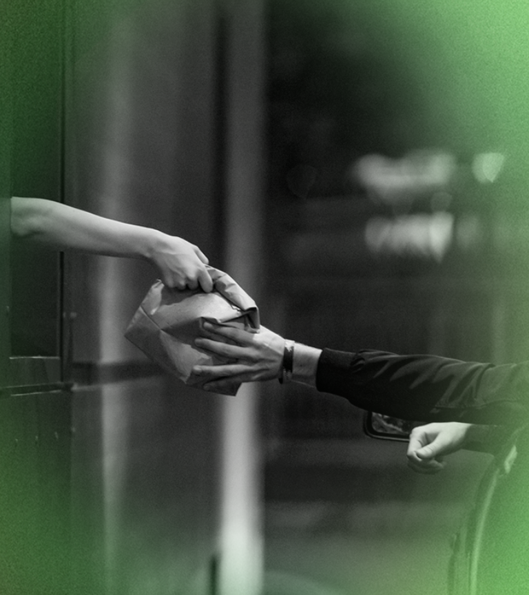 Hands passing a folded paper object, possibly a package, outdoors.