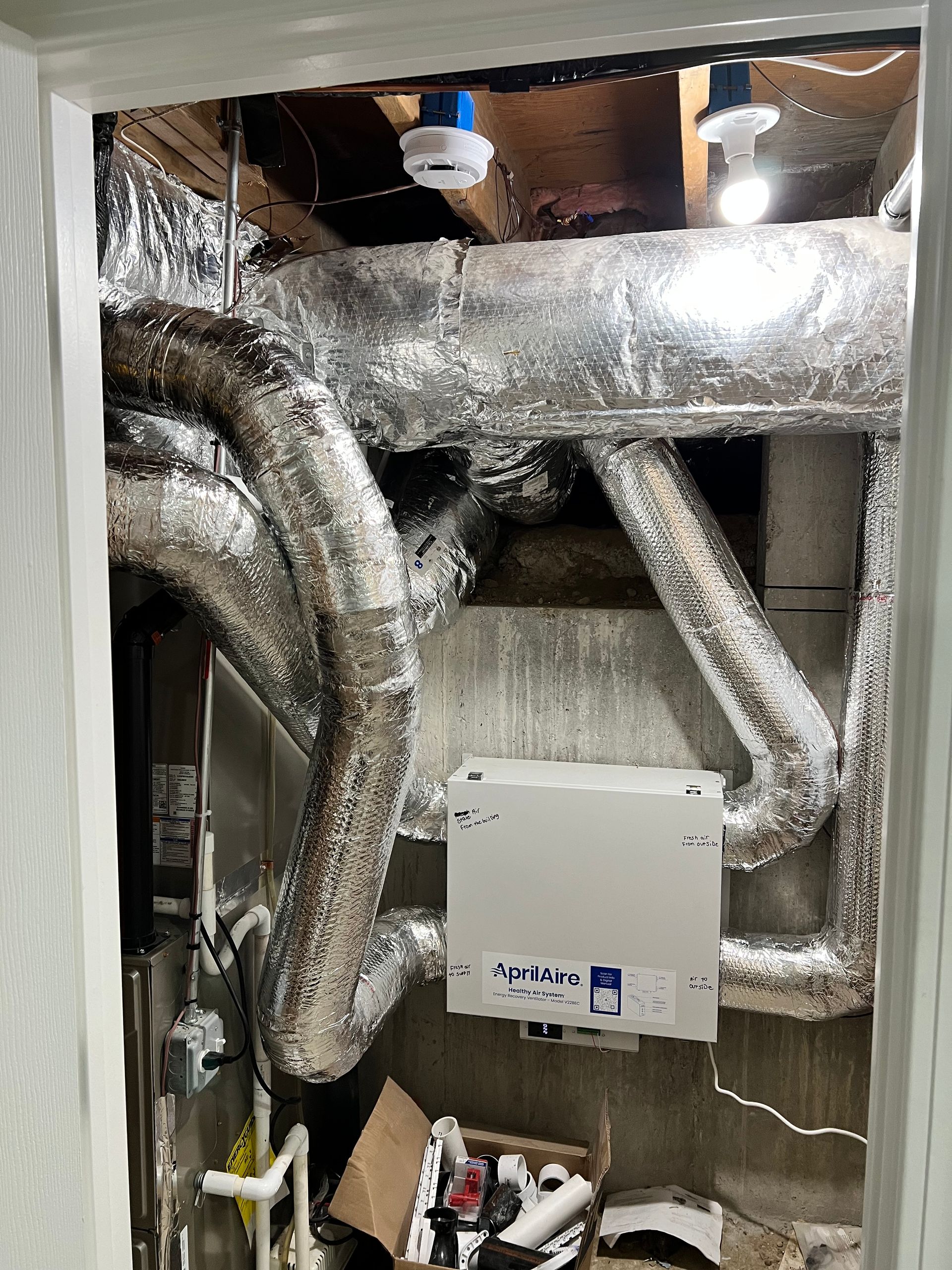 A cramped utility room with ductwork, HVAC unit, and exposed pipes. Silver insulation and a white box.