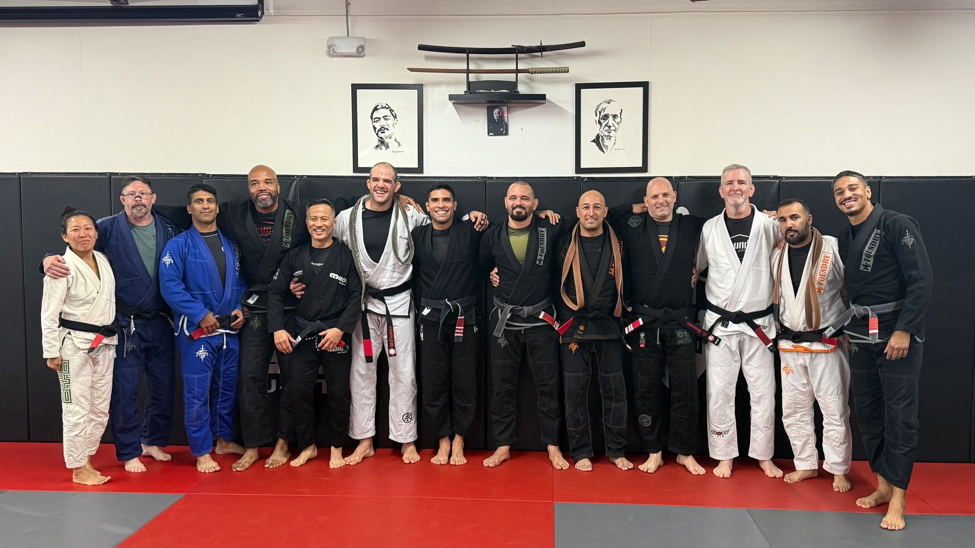 Group of people in martial arts attire posing for a photo in a dojo with artwork and mats.