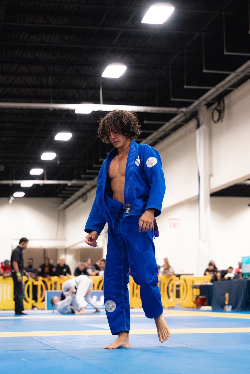 A man in a blue judo uniform stands on a mat, looking down, in a competition setting.