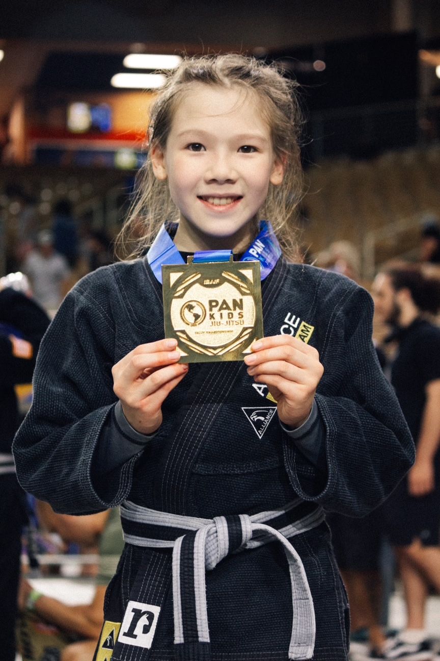 Young person in a black gi, holding a Pan Kids award, smiling.