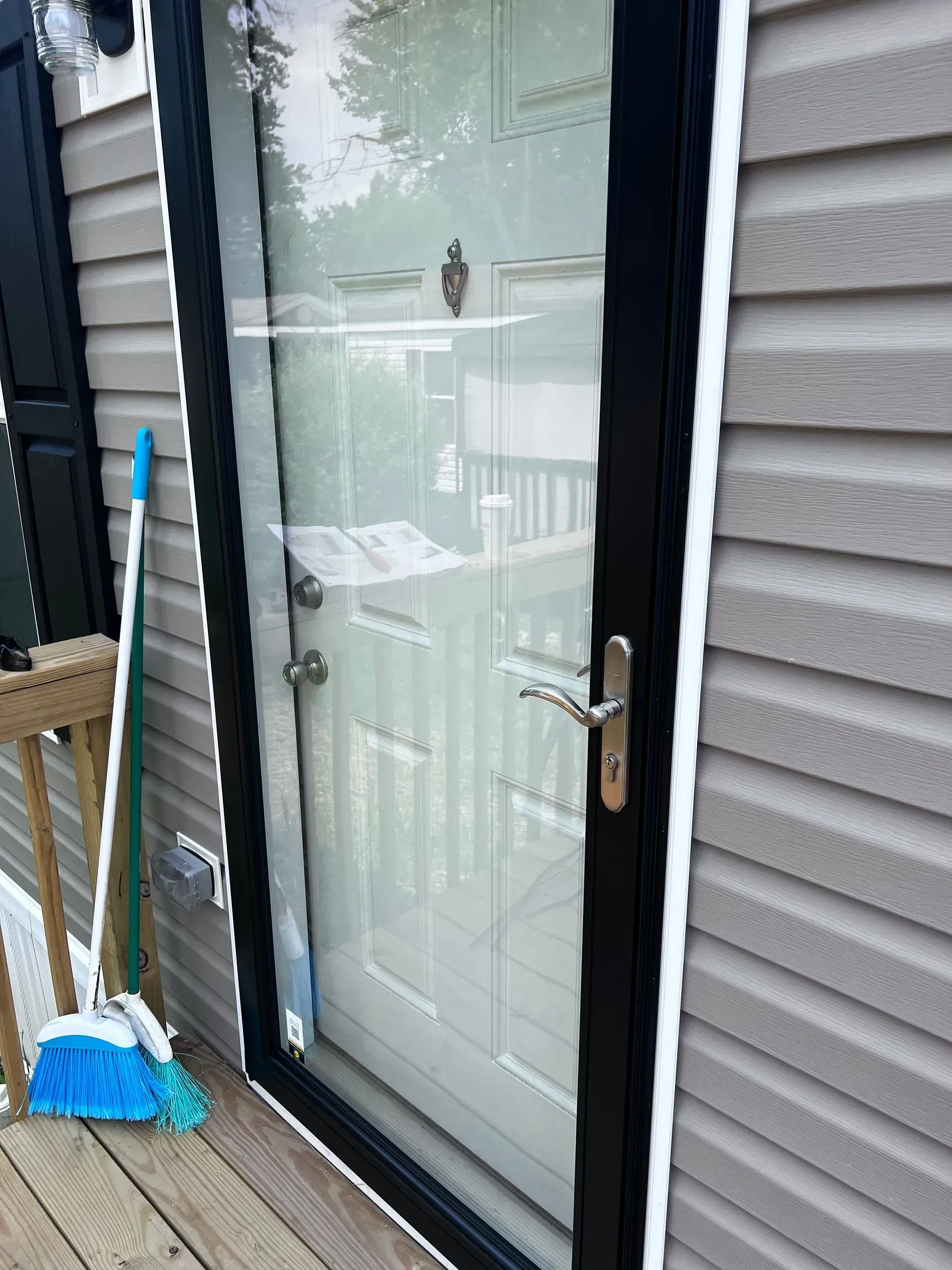A screen door, black frame, with a silver handle. A blue broom leans against the door, beside gray siding.