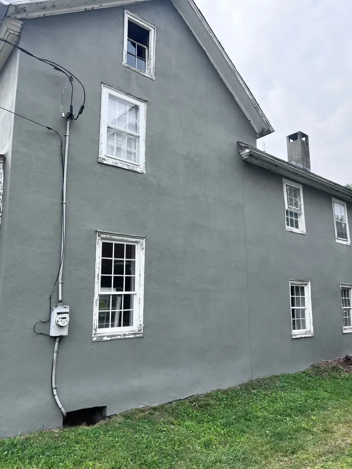 Gray stucco house with white-framed windows, electrical conduit, and a small yard.