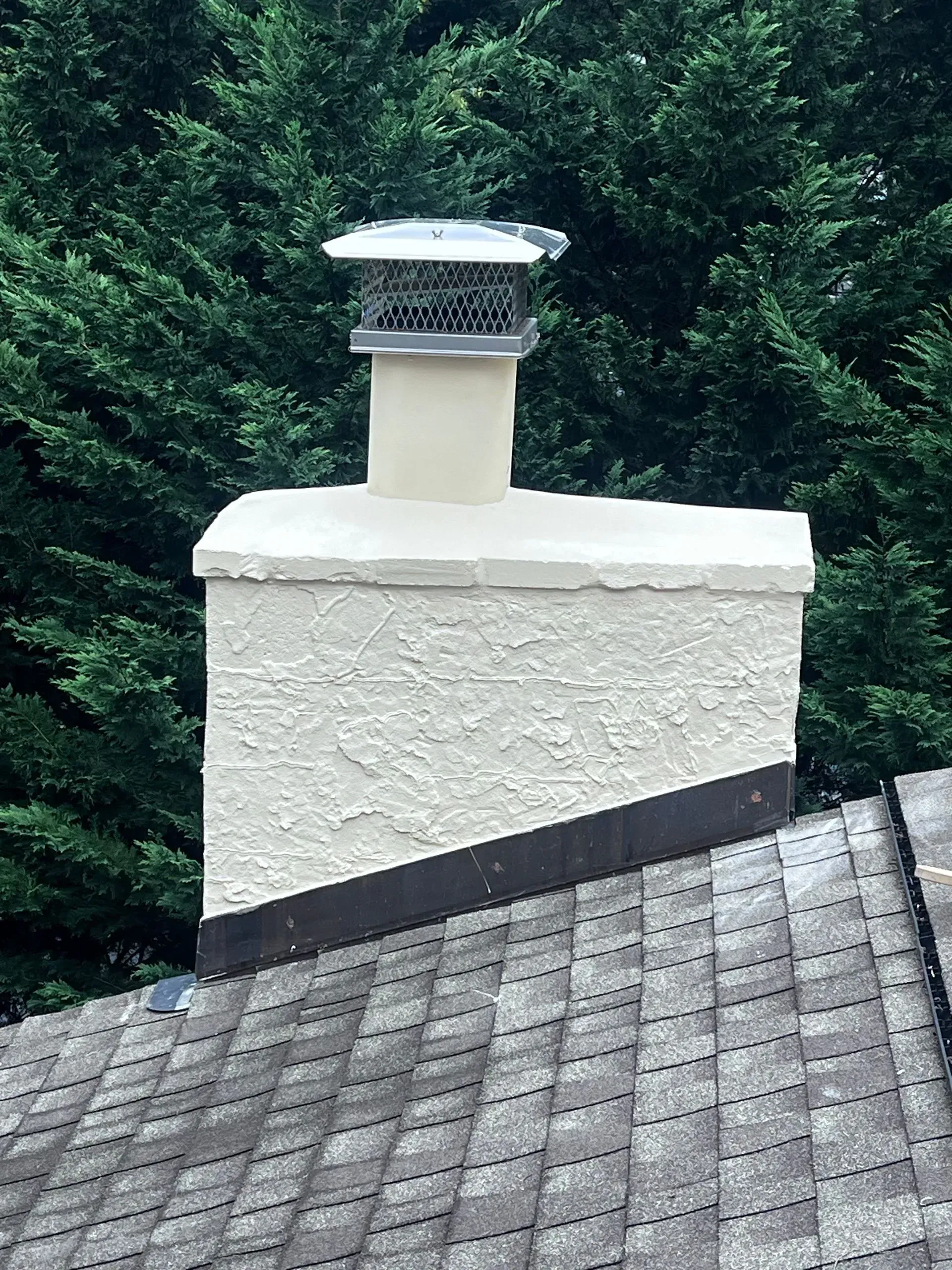 Chimney with cap on rooftop against a backdrop of green trees.