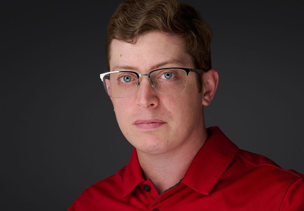 A young man wearing glasses and a red shirt is looking at the camera.