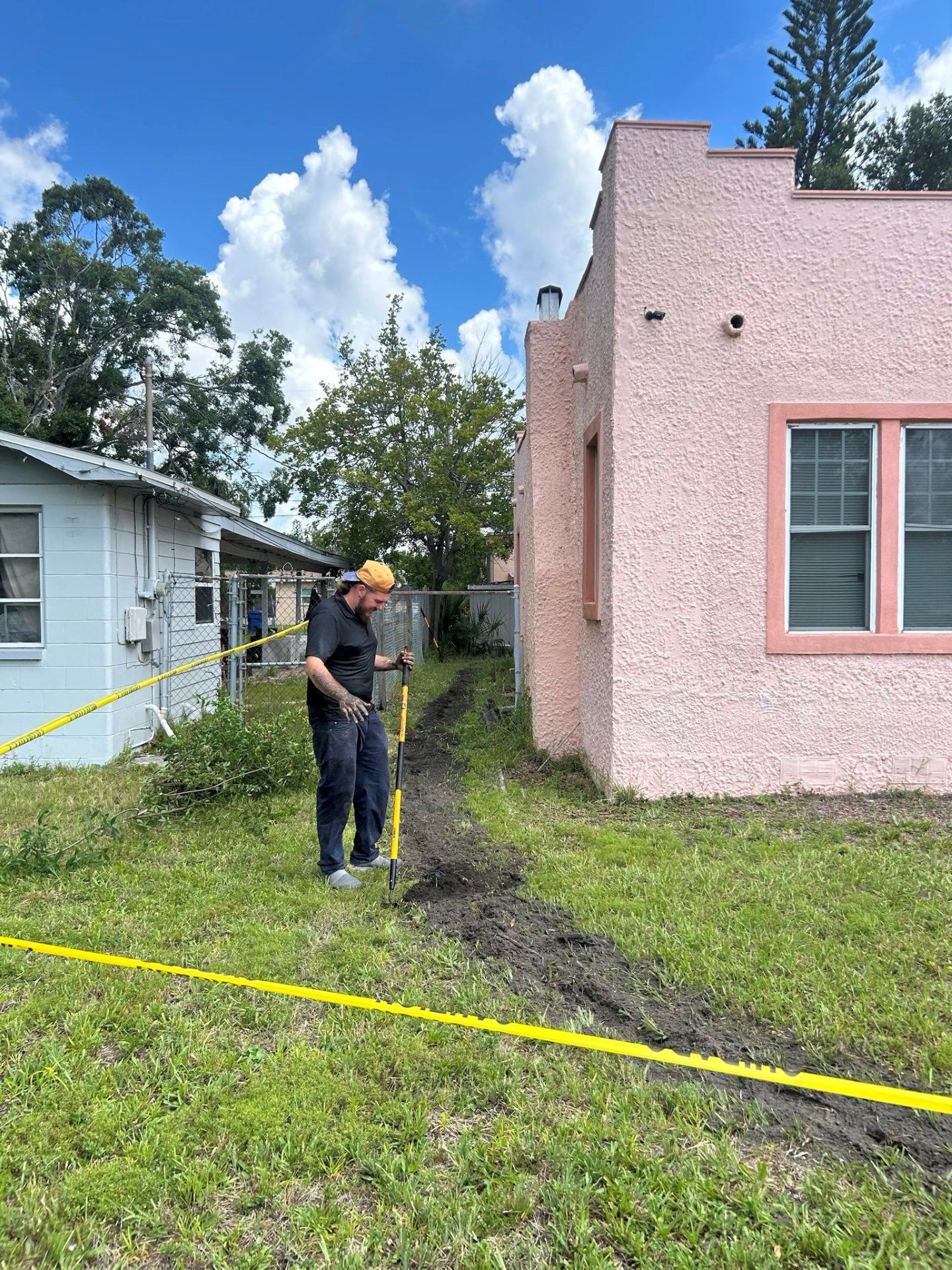Man digging in dirt near a pink building; yellow caution tape is present.