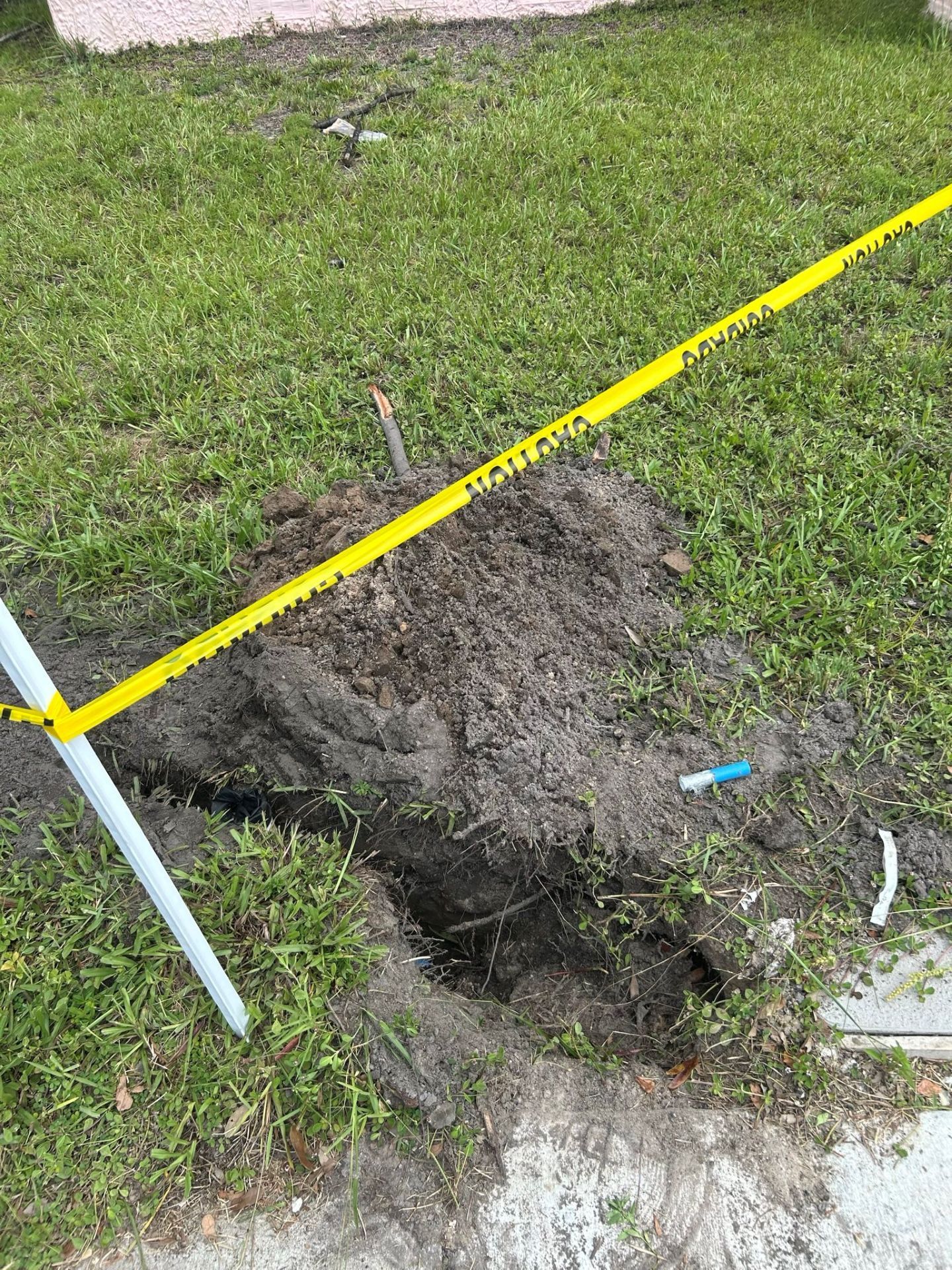 Hole in grassy lawn with yellow caution tape, near a sidewalk.