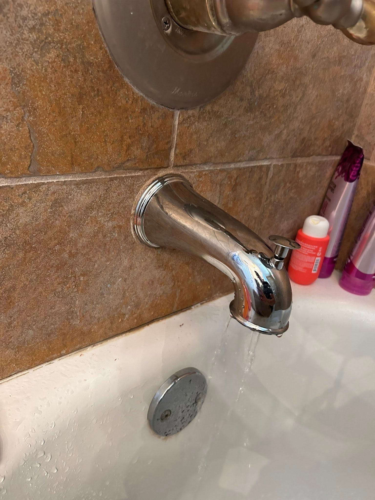 Chrome bathtub faucet with dripping water. Brown tile wall, white tub with soap.