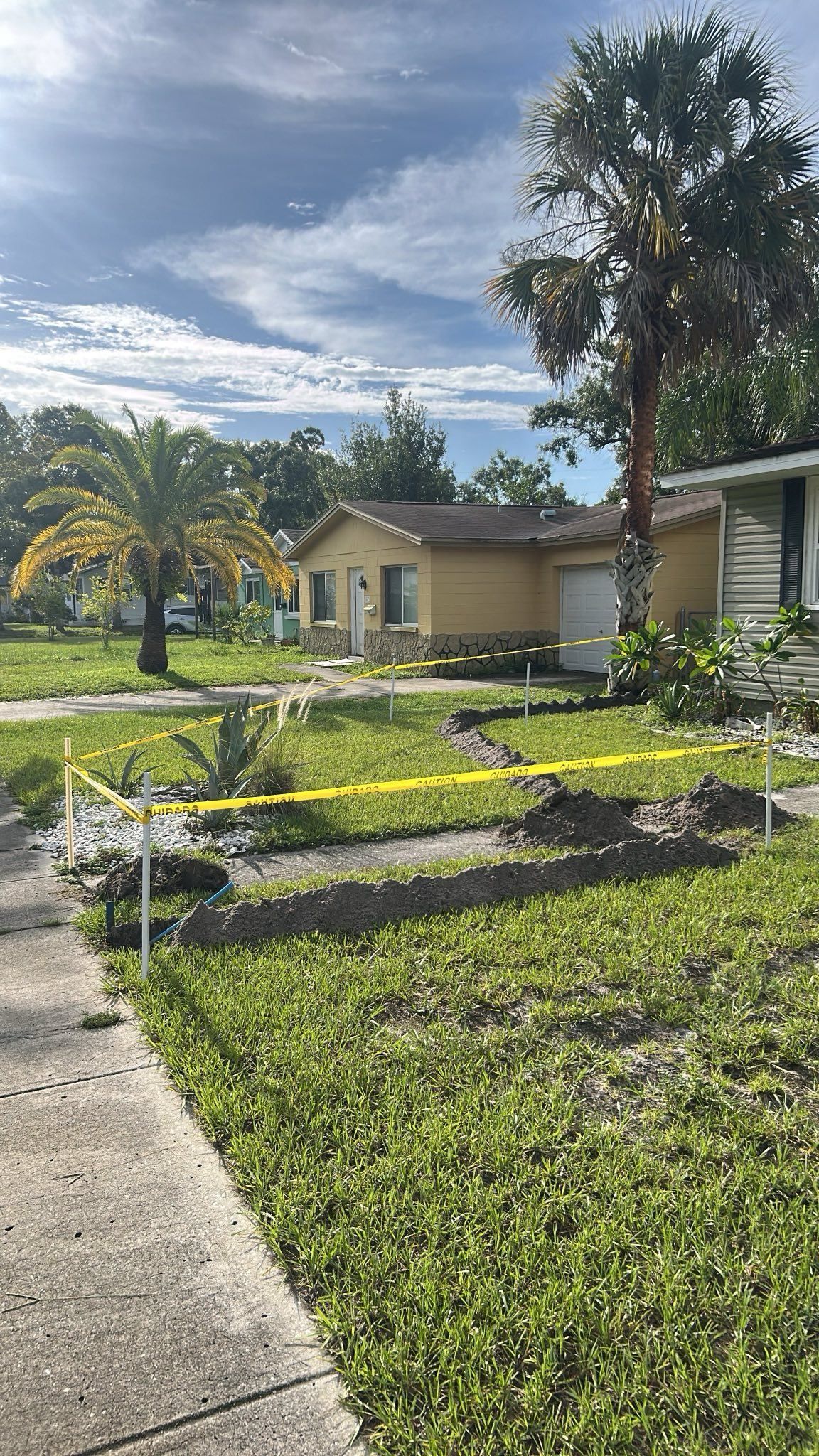 Yellow house with a damaged yard, marked with caution tape, under a partly cloudy sky.