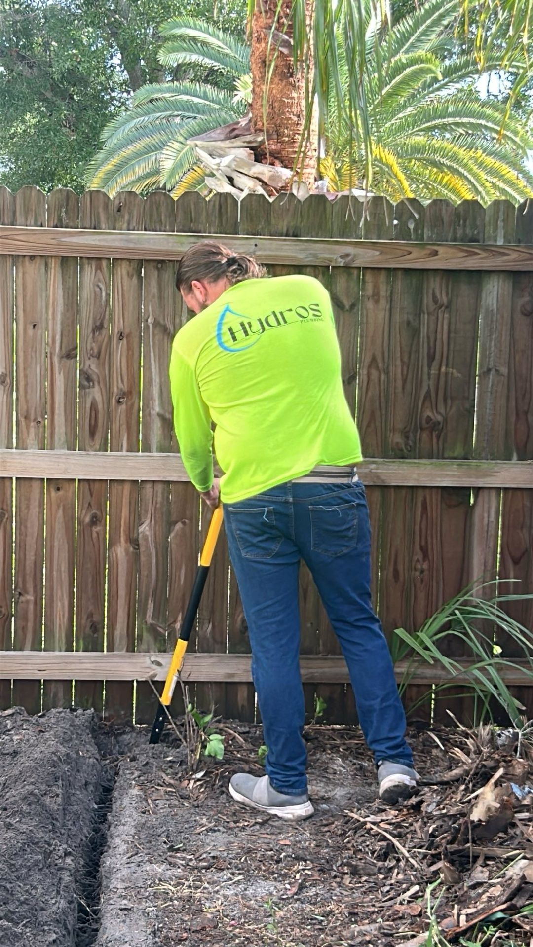 A person in a neon green shirt digs in a trench next to a wooden fence.