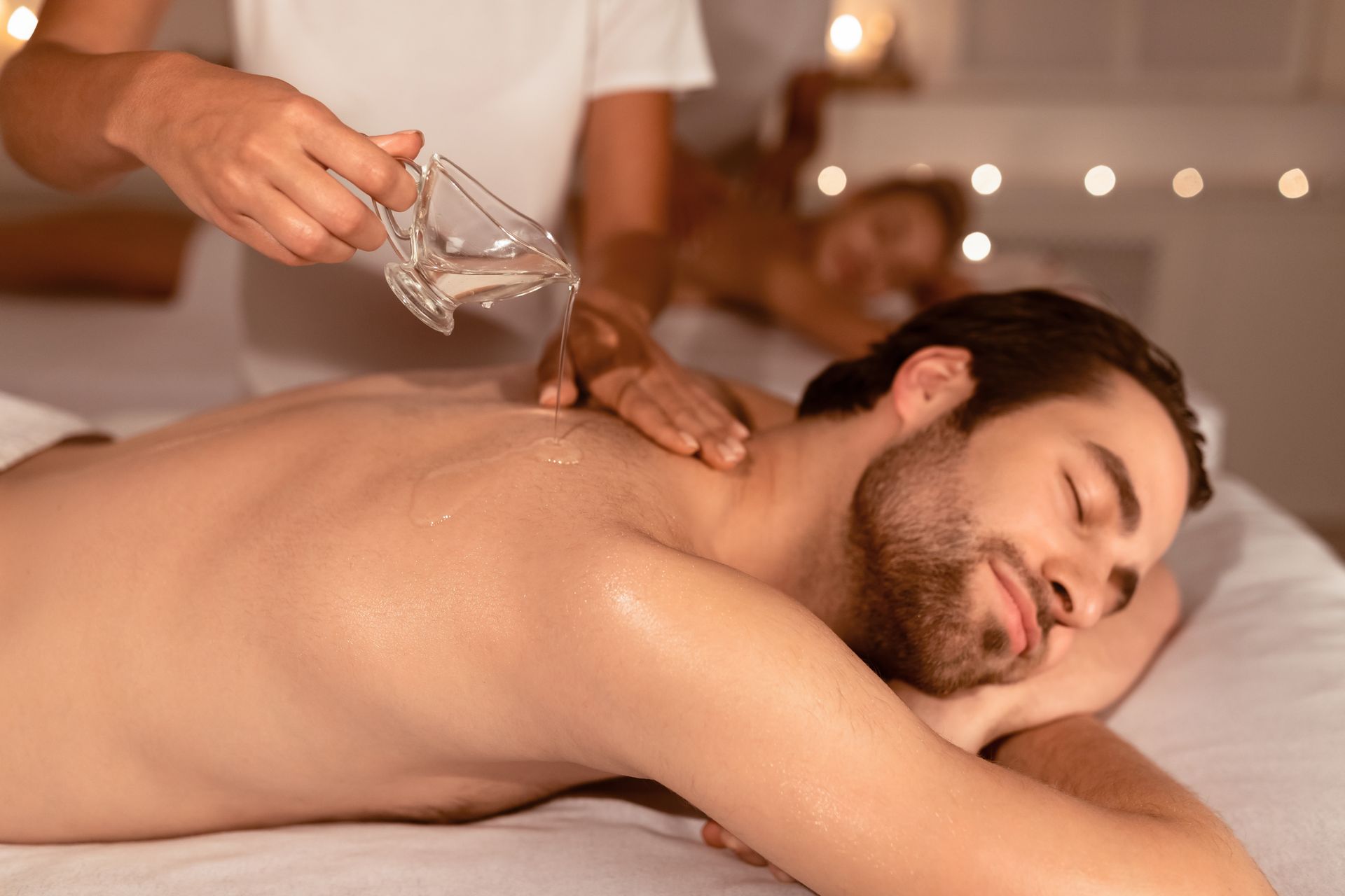 Spa therapist pouring oil on a man receiving a back massage in a warm, candlelit room