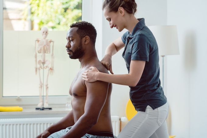 Physical therapist massaging a seated patient’s shoulder in a bright clinic room.