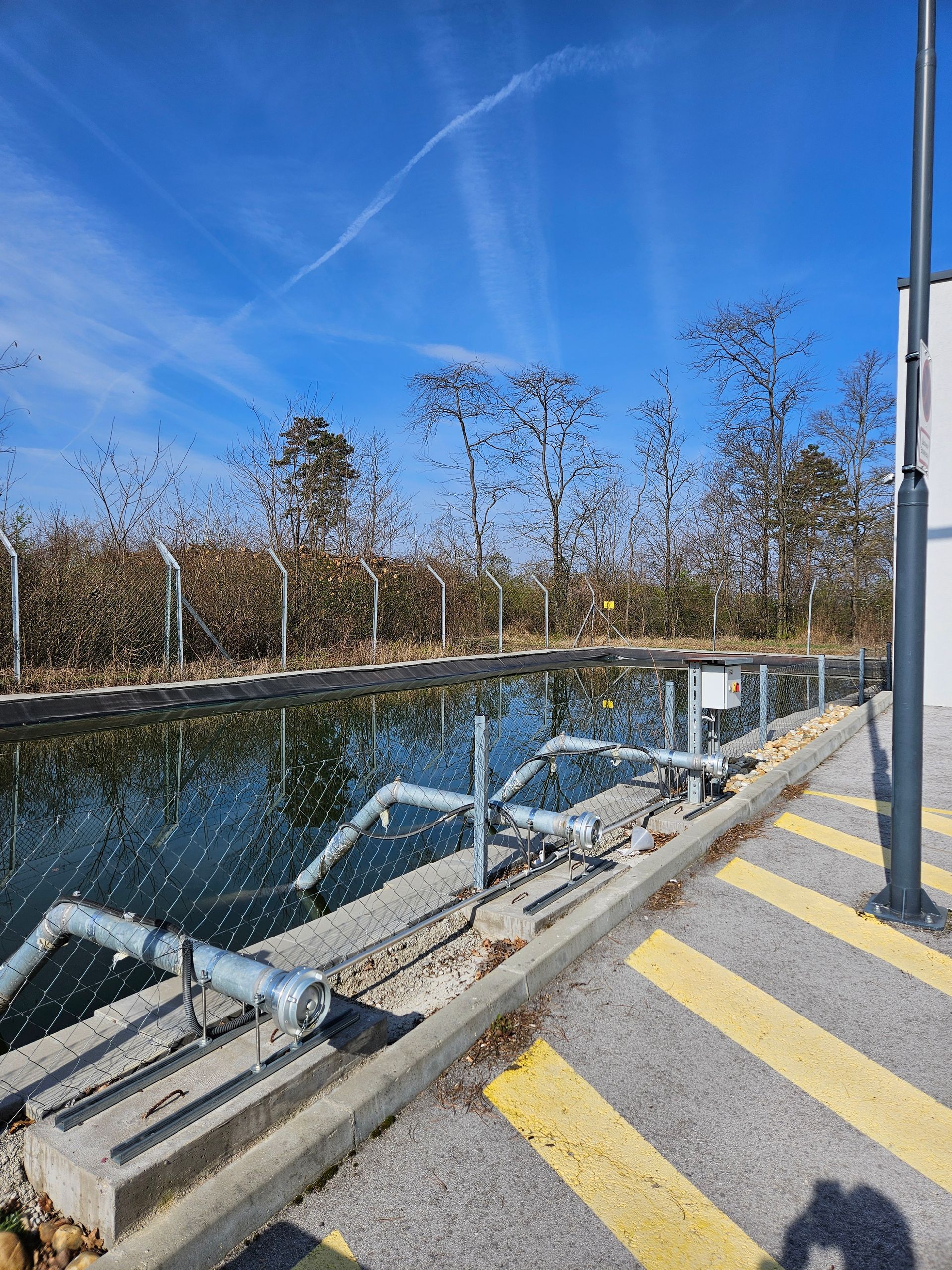 Wasseraufbereitungsanlage mit einem reflektierenden Becken, Rohren und Bäumen unter blauem Himmel.