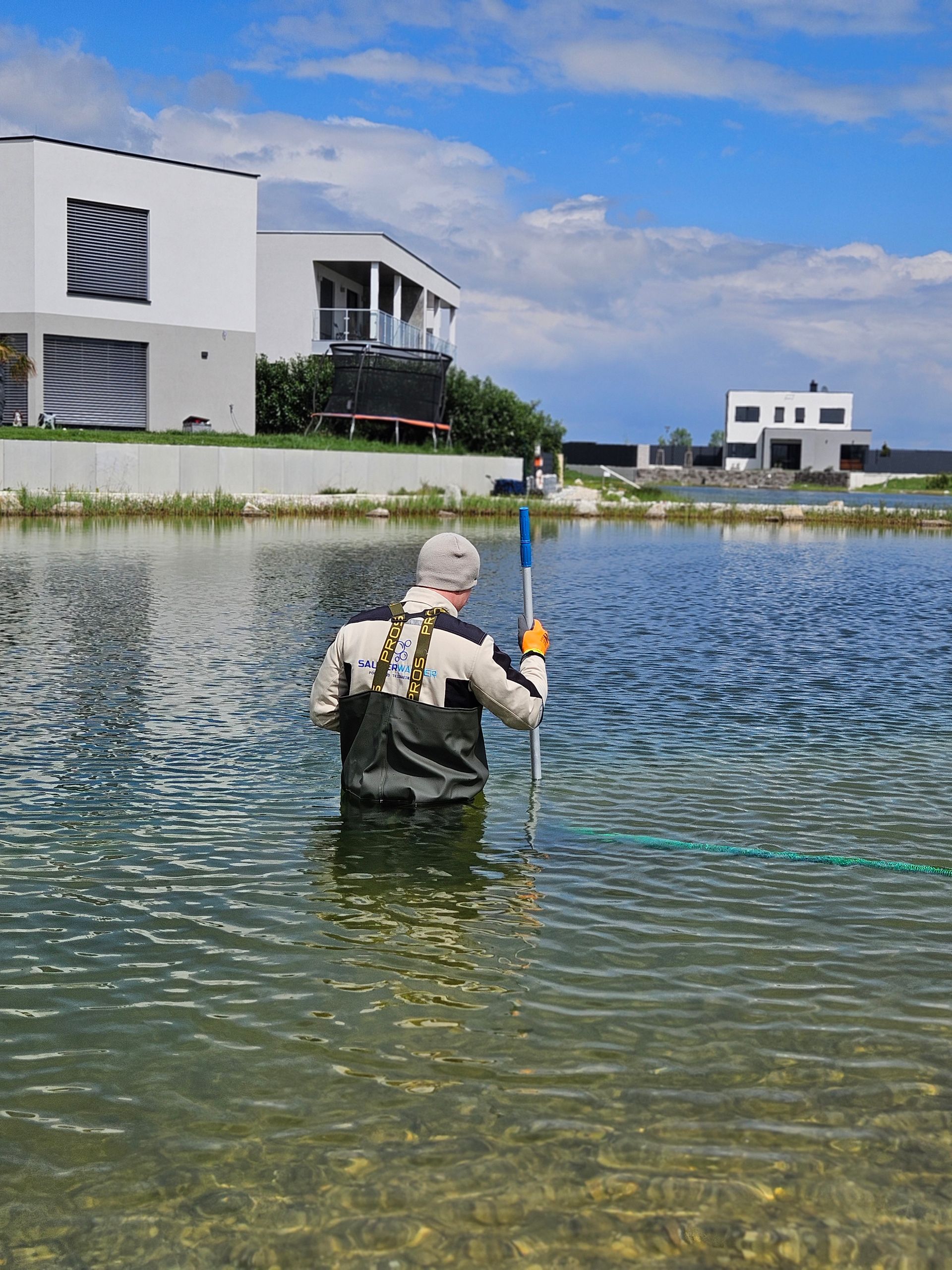 Mann in Wathose im See, hält eine Angelrute. Moderne Häuser im Hintergrund, blauer Himmel.