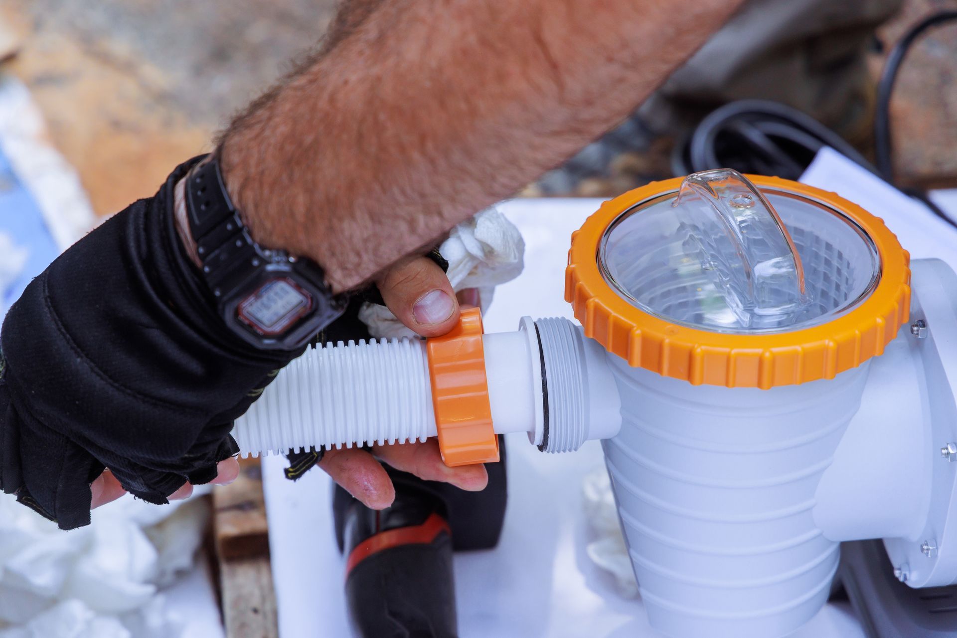 Eine Person verbindet einen weißen gerippten Schlauch mithilfe eines orangefarbenen Anschlussstücks mit einer Poolpumpe.