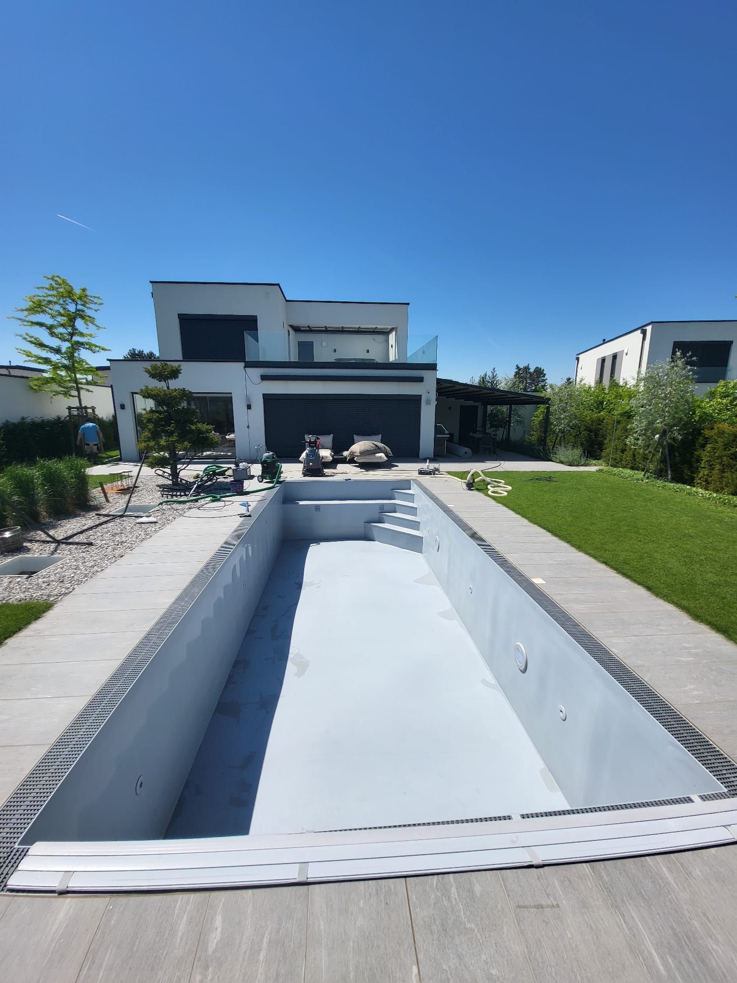 Ein leerer, rechteckiger Pool mit Treppe vor einem modernen weißen Haus unter blauem Himmel.
