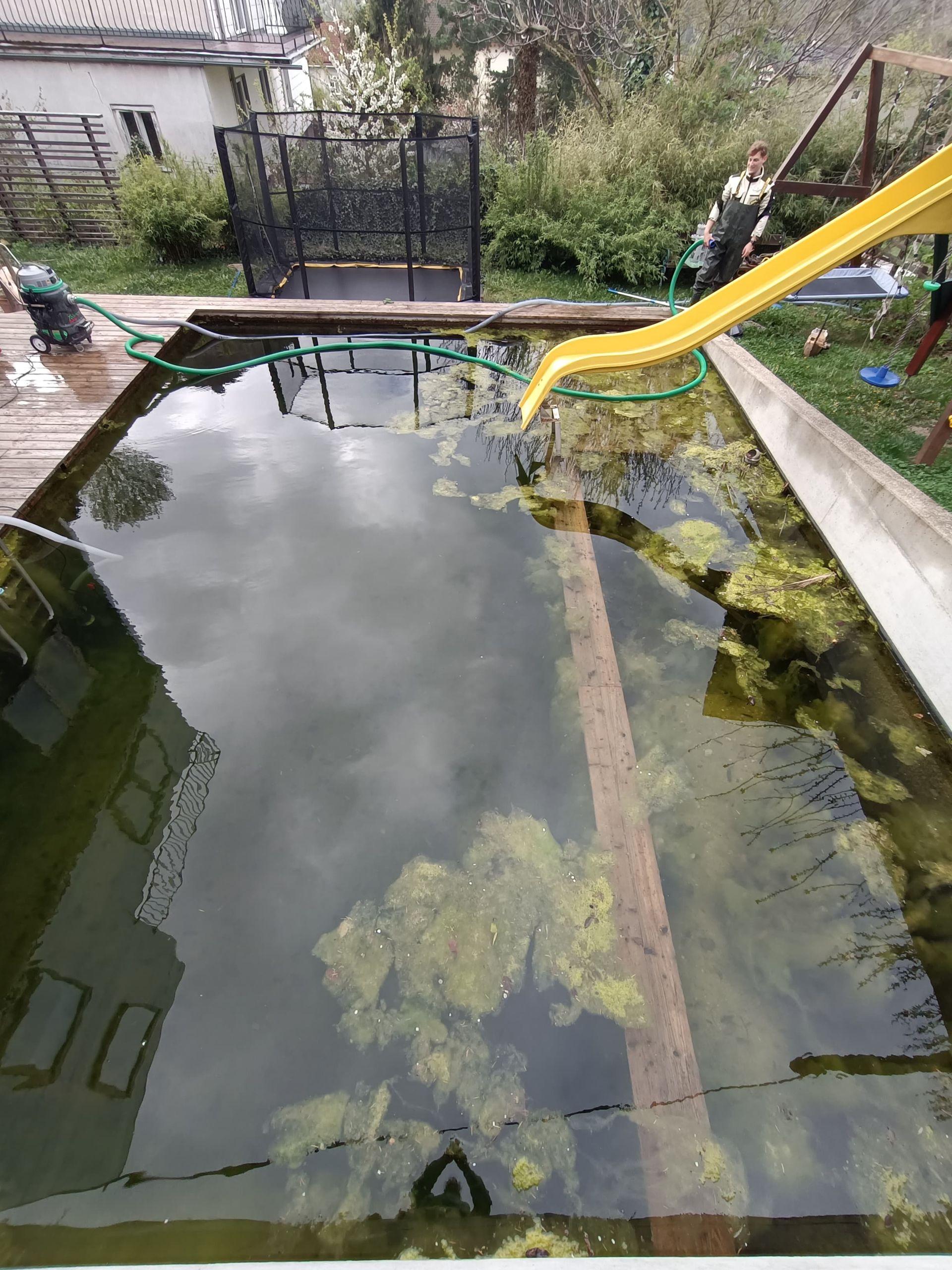 Rechteckiger Pool, dunkle Auskleidung, Holzrahmen, Wasser fließt aus einem gelben Rohr. Im Freien.