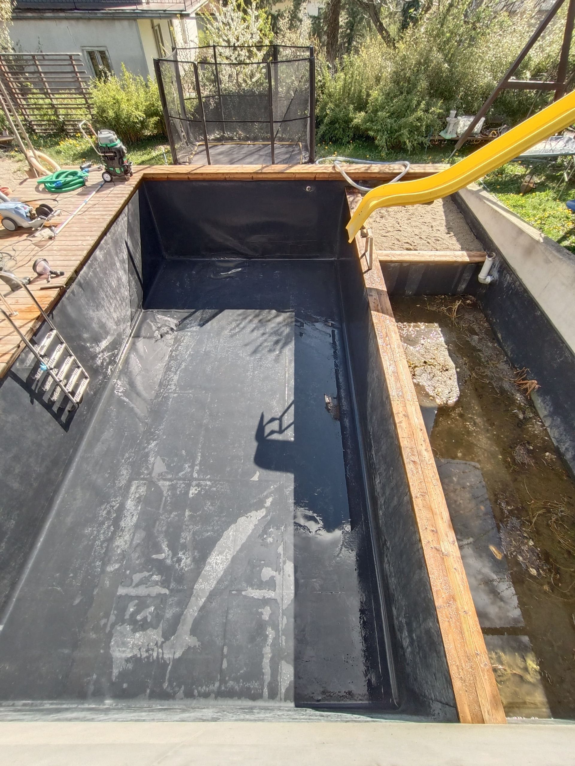 Ein mit grünen Algen gefüllter Pool, umgeben von einer Holzterrasse und einer gelben Rutsche.