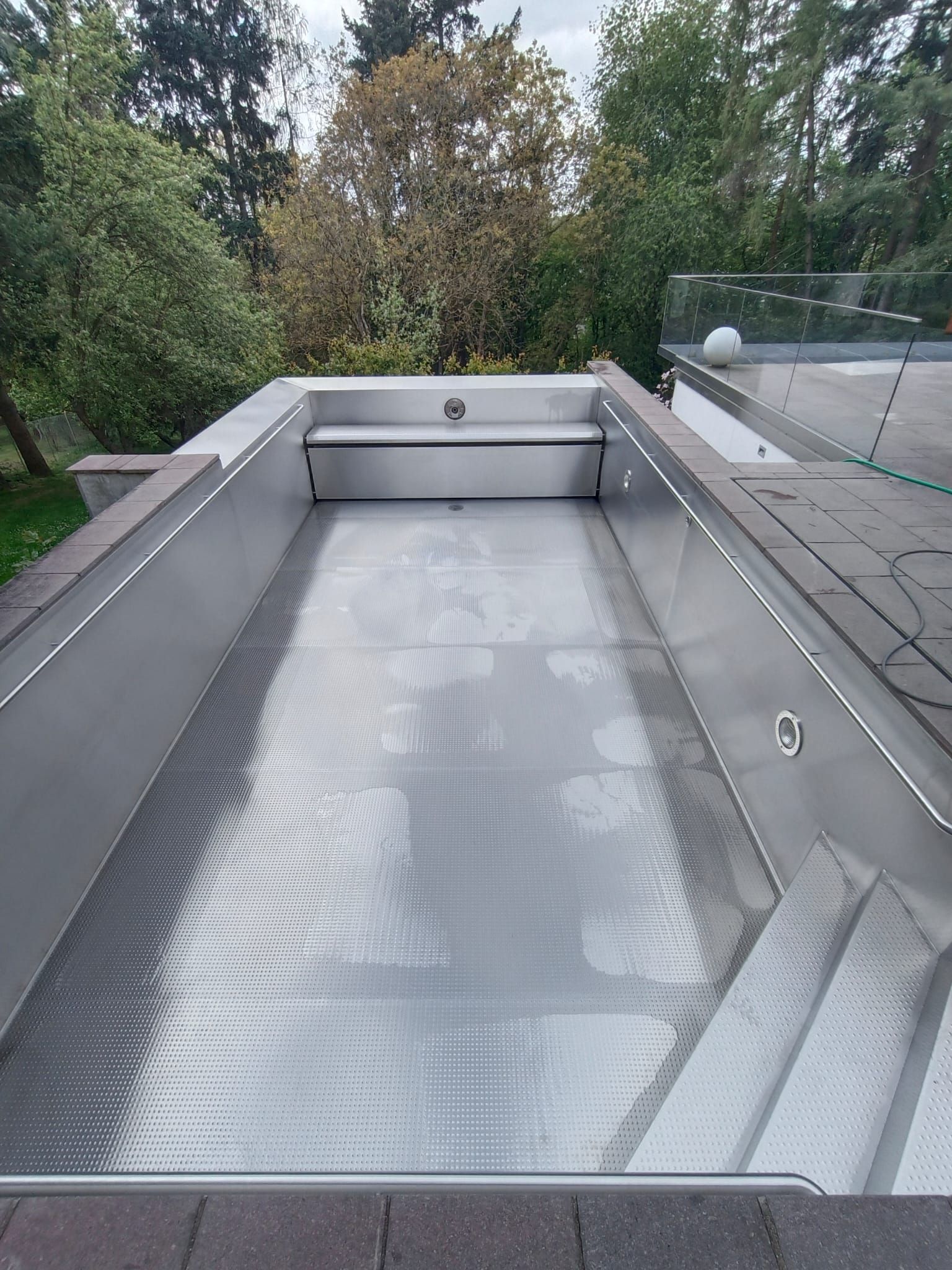 Edelstahl-Schwimmbecken mit Treppe, umgeben von einer Ziegelterrasse und Bäumen.