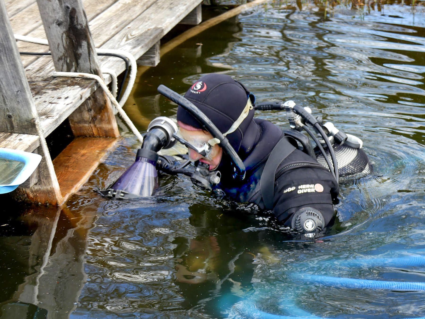Taucher in schwarzem Neoprenanzug, untergetaucht in trübem Wasser in der Nähe eines Docks, benutzt Ausrüstung.